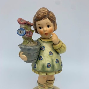May include: A porcelain figurine of a girl holding a potted plant with blue and pink flowers. The girl is wearing a green dress with white polka dots and brown shoes.