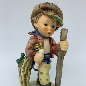 May include: A porcelain figurine of a boy wearing a brown and green hat, a red and blue plaid shirt, blue trousers, and black boots. He is carrying a stick and a bundle of sticks on his back.