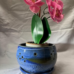 May include: A round, blue ceramic orchid pot featuring a series of small, circular ventilation holes around the upper body. The pot has a textured, mottled glaze finish and sits on a matching blue ceramic saucer, containing a pink orchid plant.