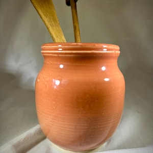 May include: A peach-colored ceramic utensil holder with a rounded shape and a light-colored base. Two wooden spoons are placed inside the holder. The holder has a glossy finish and is likely handmade. Kitchen decor.