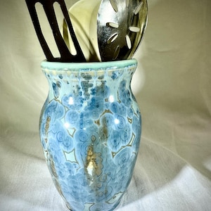 May include: A ceramic utensil holder with a blue and white floral pattern, holding a black spatula, a wooden spoon, and a slotted metal spoon. The holder has a light green rim.