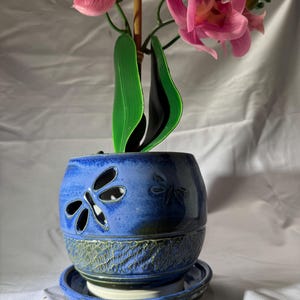 May include: A round, blue ceramic planter with a matching saucer, featuring cut-out dragonfly designs on the upper section and a textured, mottled green and blue finish on the lower base, containing a pink orchid with green leaves.