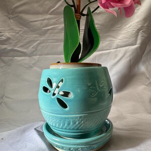 May include: A turquoise ceramic planter with a matching saucer, featuring cut-out dragonfly designs on the upper body and a textured pattern on the lower section. A faux pink orchid plant with green leaves is potted inside the round container.
