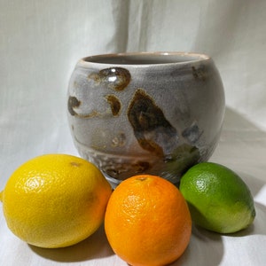 May include: A ceramic bowl with a white glaze and brown and gray marbling. The bowl is sitting on a white surface with three pieces of fruit: a lemon, an orange, and a lime.
