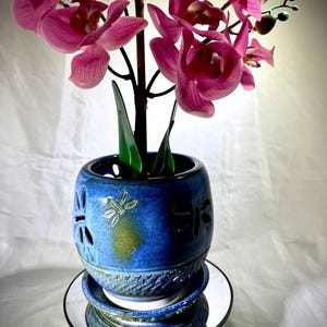 May include: A blue ceramic planter with dragonfly cutouts, holding a pink orchid. The planter sits on a mirrored base. The orchid has vibrant pink petals and green leaves. The planter has a glazed finish.