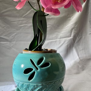 May include: A turquoise ceramic planter with a dragonfly cutout design and a matching saucer. The planter features a textured base and holds a potted artificial orchid with vibrant pink petals and dark green leaves, topped with small black stones.