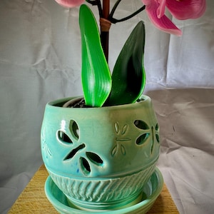 May include: A turquoise ceramic planter with dragonfly cutouts and a matching saucer. The planter holds a plant with green leaves and pink orchid flowers. The planter sits on a wooden surface.