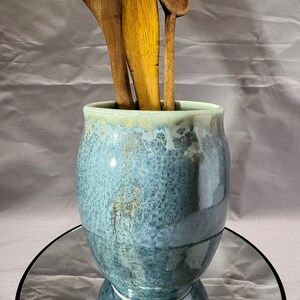 May include: A cylindrical ceramic utensil holder with a mottled blue and grey glaze featuring a drip effect near the rim. Three wooden kitchen spoons of varying sizes are placed inside the container, which has a smooth, glossy finish.