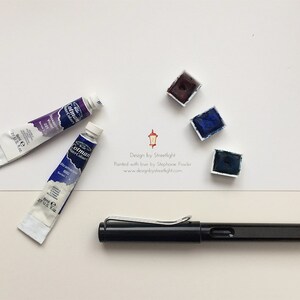 May include: A black fountain pen, two tubes of watercolor paint, and three watercolor paint pans. The paint pans are in shades of blue and purple. The text on the white card reads "Design by Streetlight. Painted with love by Stephanie Fowler. www.designbystreetlight.com"