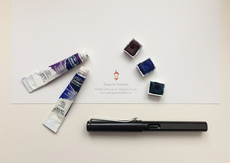 May include: A black fountain pen, two tubes of watercolor paint, and three watercolor paint pans. The paint pans are in shades of blue and purple. The text on the white card reads "Design by Streetlight. Painted with love by Stephanie Fowler. www.designbystreetlight.com"