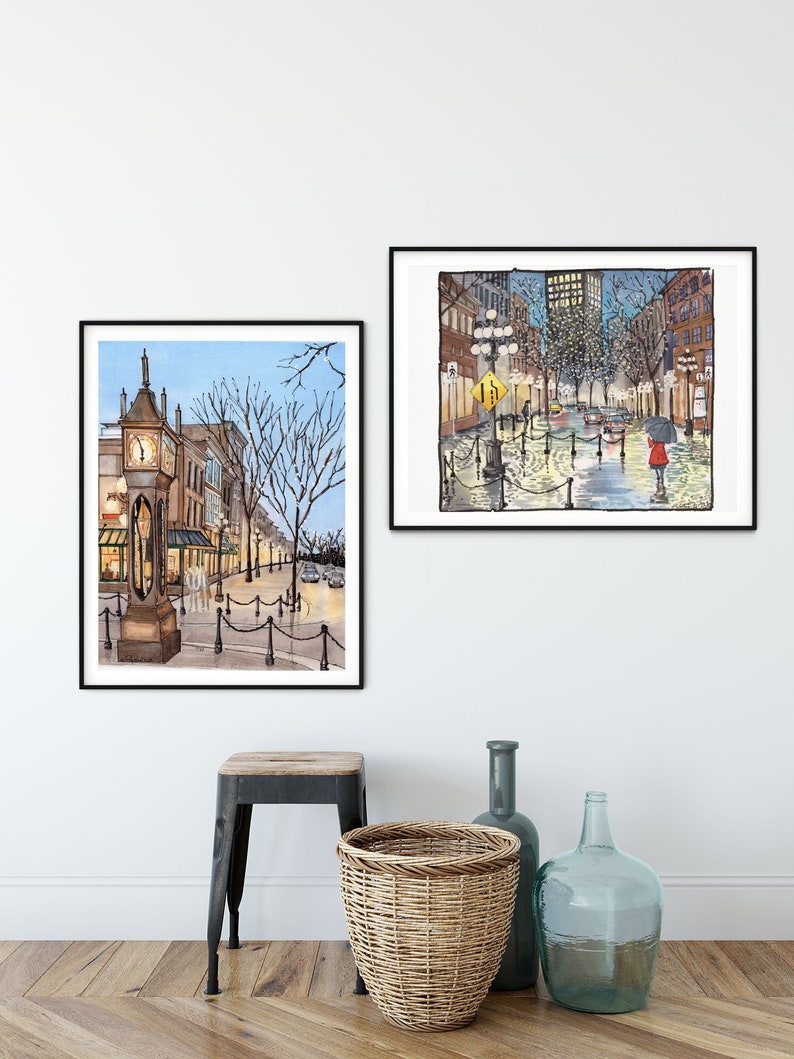 May include: Two framed watercolour paintings of city streets. The painting on the left shows a clock tower and buildings with a blue sky. The painting on the right shows a street with buildings and streetlights at night with a person walking with an umbrella.
