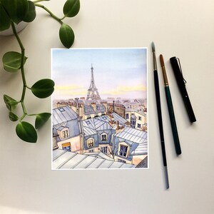 May include: A watercolor painting of a Parisian cityscape with the Eiffel Tower in the distance. The painting features a view from a rooftop looking out over the city, with many buildings and rooftops visible. The sky is a light blue with a hint of pink in the distance.