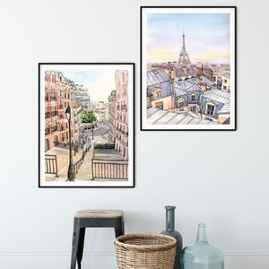 May include: Two framed watercolor paintings of Parisian street scenes. The first painting shows a view of a street with buildings on either side and a lamppost in the center. The second painting shows a view of rooftops with the Eiffel Tower in the distance.