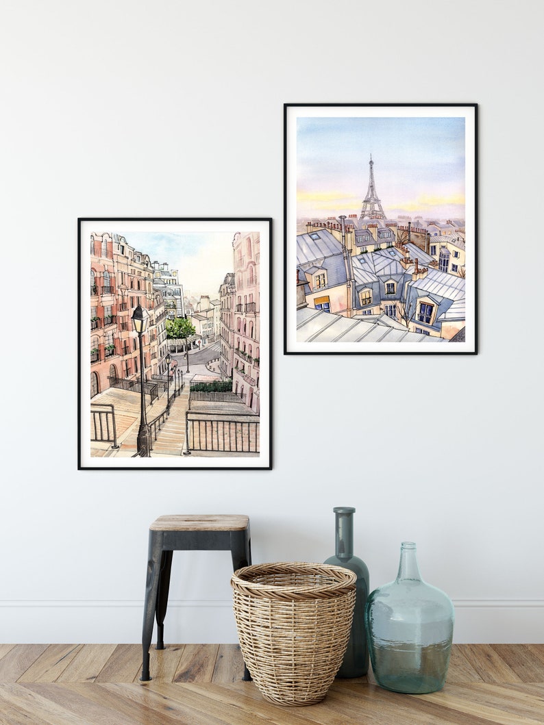 May include: Two framed watercolor paintings of Parisian street scenes. The first painting shows a view of a street with buildings on either side and a lamppost in the center. The second painting shows a view of rooftops with the Eiffel Tower in the distance.