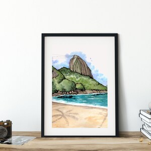 May include: A framed watercolour painting depicting a beach scene with a large, rocky mountain in the background. The water is blue and the sand is light brown. There are palm trees in the foreground.