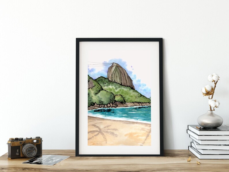 May include: A framed watercolour painting depicting a beach scene with a large, rocky mountain in the background. The water is blue and the sand is light brown. There are palm trees in the foreground.