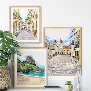 May include: Three framed watercolor paintings depicting scenes from Rio de Janeiro, Brazil. The first painting shows a narrow street lined with colorful buildings. The second painting shows a cobblestone square with a church in the background. The third painting shows a beach with Sugarloaf Mountain in the distance.