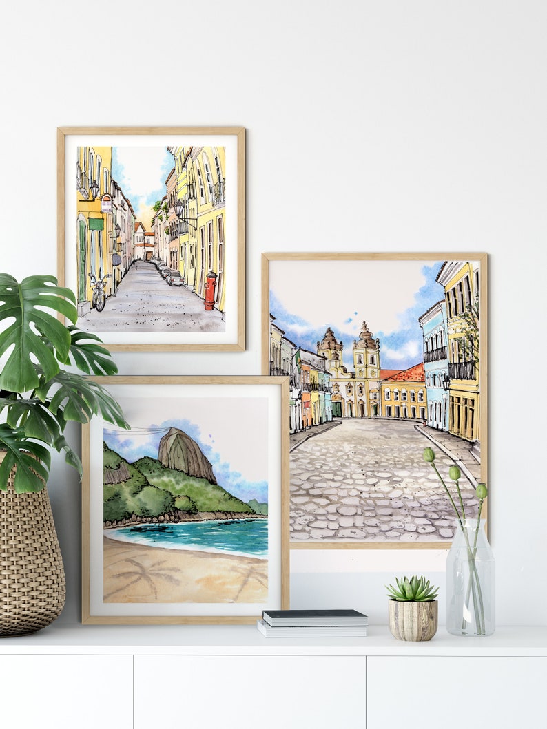 May include: Three framed watercolor paintings depicting scenes from Rio de Janeiro, Brazil. The first painting shows a narrow street lined with colorful buildings. The second painting shows a cobblestone square with a church in the background. The third painting shows a beach with Sugarloaf Mountain in the distance.