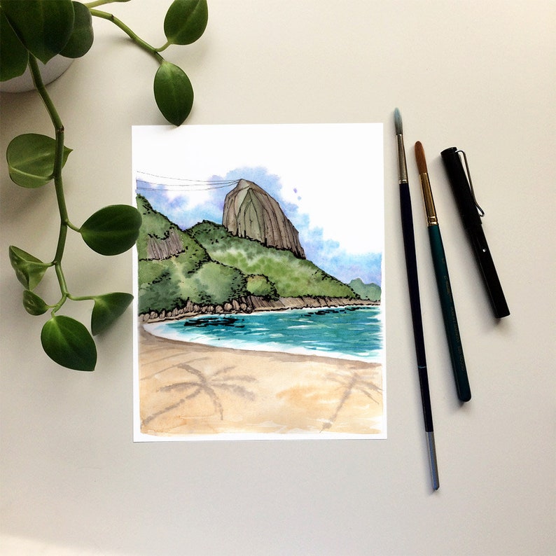 May include: Watercolor painting of a beach scene with a mountain in the background. The beach is sandy with palm tree shadows and the water is blue. The mountain is green and brown.
