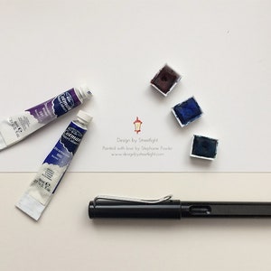 May include: A black fountain pen, two tubes of watercolour paint, and three watercolour paint pans. The paint pans are in shades of blue and purple. The text on the white card reads "Design by Streetlight. Painted with love by Stephanie Fowler. www.designbystreetlight.com"