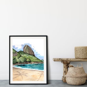 May include: A framed watercolor painting of a beach scene with a mountain in the background. The painting is in a black frame and is hanging on a white wall. There is a wooden bench with two woven baskets in front of the painting.