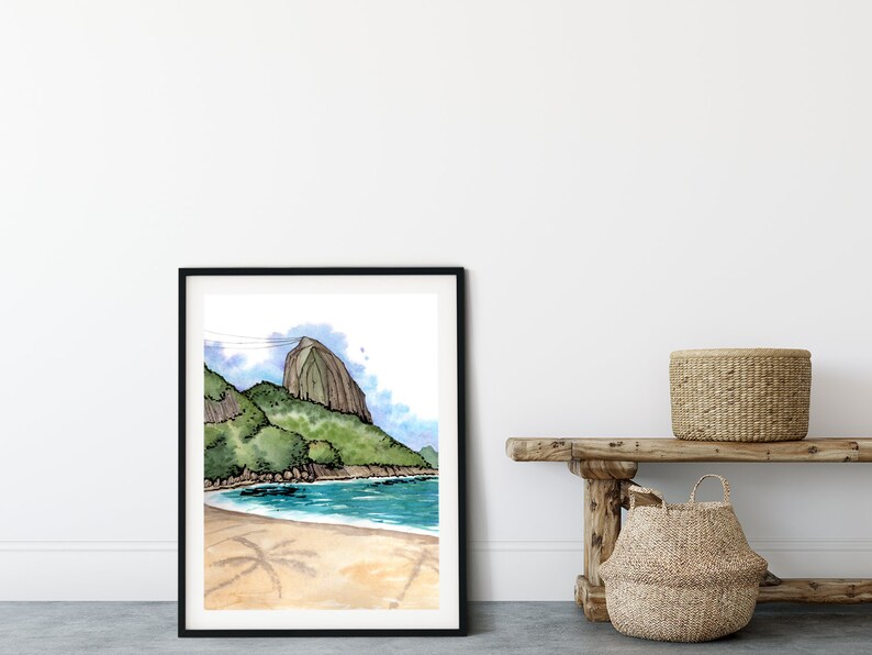 May include: A framed watercolor painting of a beach scene with a mountain in the background. The painting is in a black frame and is hanging on a white wall. There is a wooden bench with two woven baskets in front of the painting.