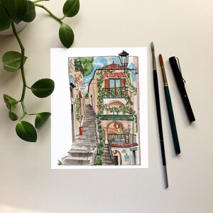 May include: Watercolor painting of a building with a red roof and a green awning. The building has a balcony with a wrought iron railing and a window with a view of the street below. The building is located on a hill and there are stairs leading up to it. The painting is done in a realistic style and the colors are bright and vibrant.