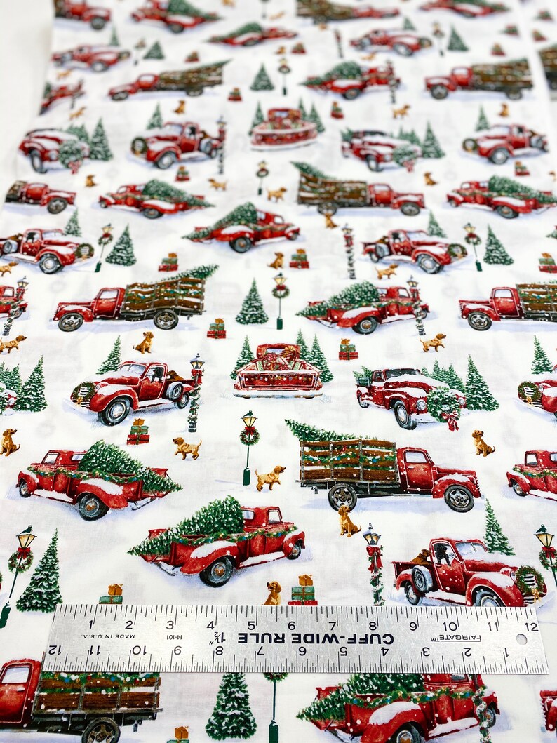 K&ouml;nnte beinhalten: Wei&szlig;er Stoff mit einem sich wiederholenden Muster aus roten Oldtimern, die Weihnachtsb&auml;ume, Schnee und kleine braune Hunde transportieren. Gr&uuml;ne Weihnachtsb&auml;ume und Stra&szlig;enlaternen sind ebenfalls abgebildet. Das Muster ist etwa 30 cm breit.