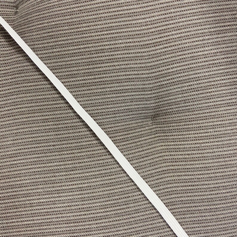 May include: A diagonal view of a white elastic band against a textured, brown fabric background. The elastic band is thin and appears to be made of woven material. The fabric has a woven pattern.
