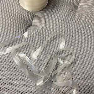 May include: A roll of clear elastic ribbon on a gray fabric surface. The ribbon is translucent and has a slight sheen.