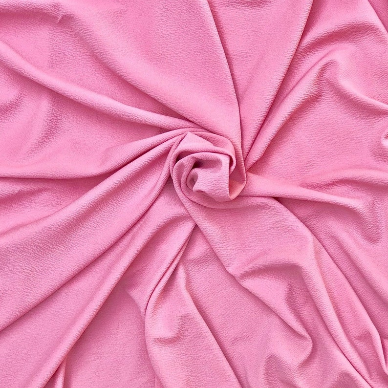 May include: A solid light pink fabric with a distinct textured surface. The material is gathered and draped in a central spiral pattern, creating soft folds and shadows that highlight the consistent color and the fine, pebbled grain of the textile.