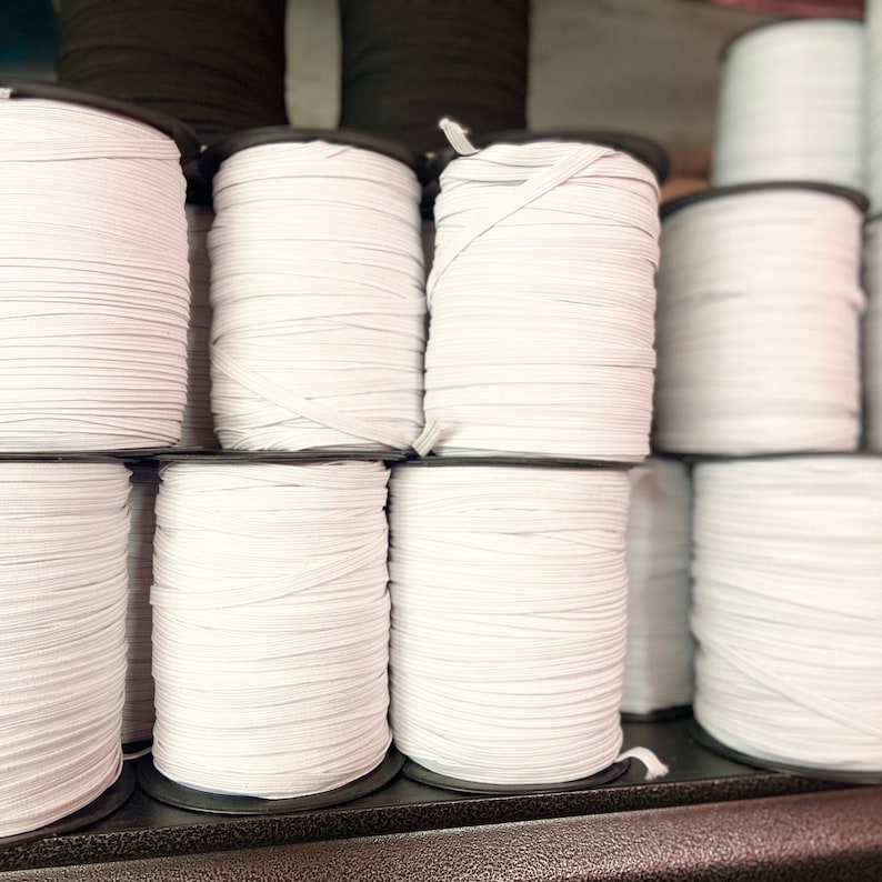 May include: A collection of white elastic cord spools, neatly stacked on a shelf. The spools are cylindrical, with the elastic wound tightly around them. The elastic appears to be flat and approximately 1/4 inch wide. A few black spools are visible in the background.