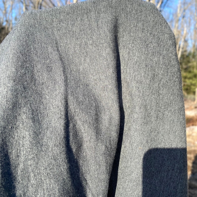 May include: A close-up shot of a gray fabric, possibly a sweatshirt or sweater. The material appears soft and textured, with subtle variations in tone. The lighting suggests an outdoor setting.