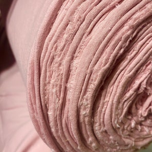 May include: A roll of soft, light pink fabric. The fabric is tightly rolled, revealing the edges of the material. The edges have a frayed, textured appearance. The fabric appears to be a knit or jersey material, suitable for clothing or crafts.