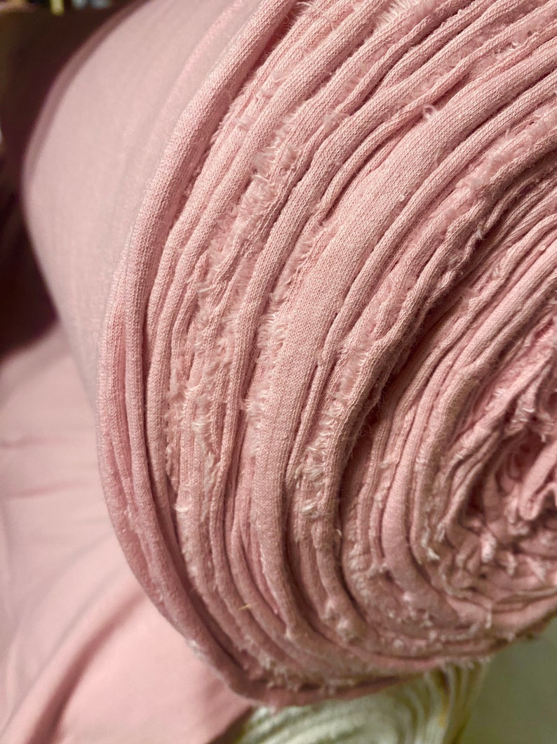 May include: A roll of soft, light pink fabric. The fabric is tightly rolled, revealing the edges of the material. The edges have a frayed, textured appearance. The fabric appears to be a knit or jersey material, suitable for clothing or crafts.