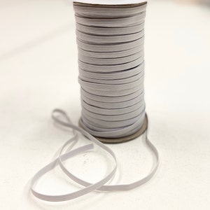 May include: A spool of light gray elastic ribbon. The elastic is tightly wound around a cardboard core. A length of the elastic ribbon is unwound and lies in front of the spool. The elastic is a flat, woven material.