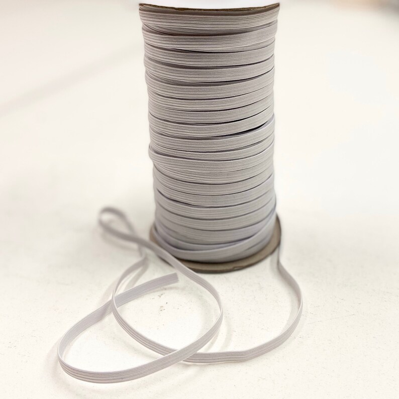 May include: A spool of light gray elastic ribbon. The elastic is tightly wound around a cardboard core. A length of the elastic ribbon is unwound and lies in front of the spool. The elastic is a flat, woven material.
