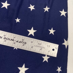 May include: A close-up of a navy blue fabric with a white star pattern. The fabric is measured with a ruler that reads "CUFF-WIDE" and has markings in inches.