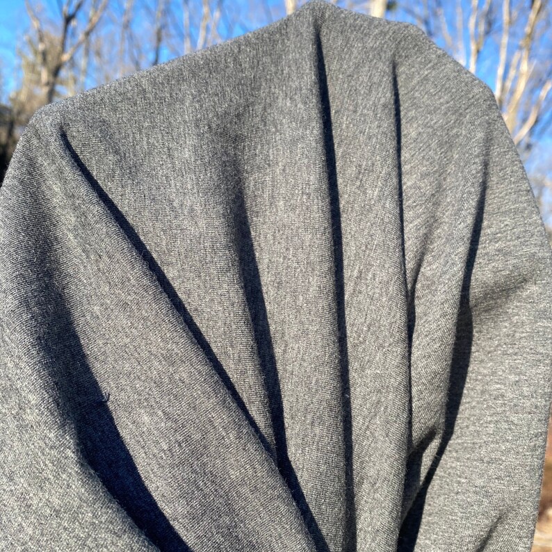 May include: Close-up of a gray fabric with visible texture and folds. The fabric is a solid, medium gray color, with shadows creating depth and dimension. The background shows a blurred view of a sunny day.
