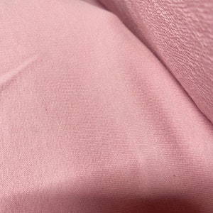 May include: Close-up of a roll of soft, pink fabric. The fabric has a textured surface with a subtle ribbed pattern. The color is a muted, dusty rose, and the material appears to be a knit or similar textile.