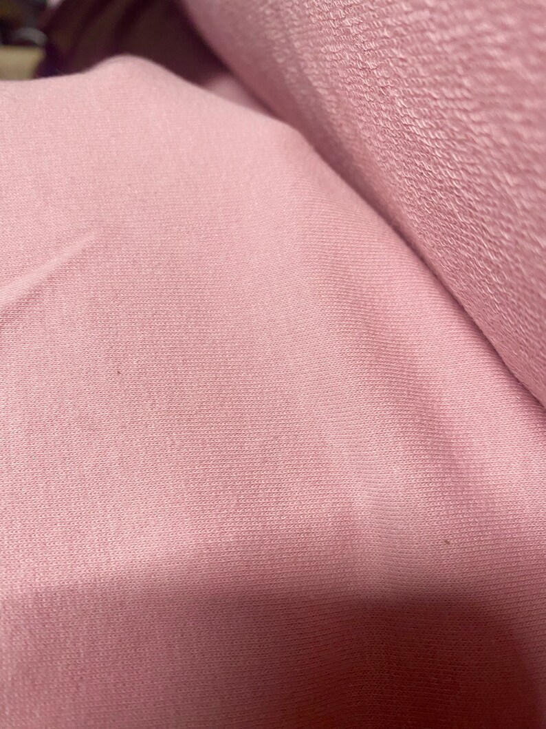 May include: Close-up of a roll of soft, pink fabric. The fabric has a textured surface with a subtle ribbed pattern. The color is a muted, dusty rose, and the material appears to be a knit or similar textile.