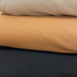 May include: A stack of three folded waffle knit fabrics in neutral colors. The top fabric is a light beige, followed by a tan and then a black fabric. The fabrics have a textured, grid-like pattern.