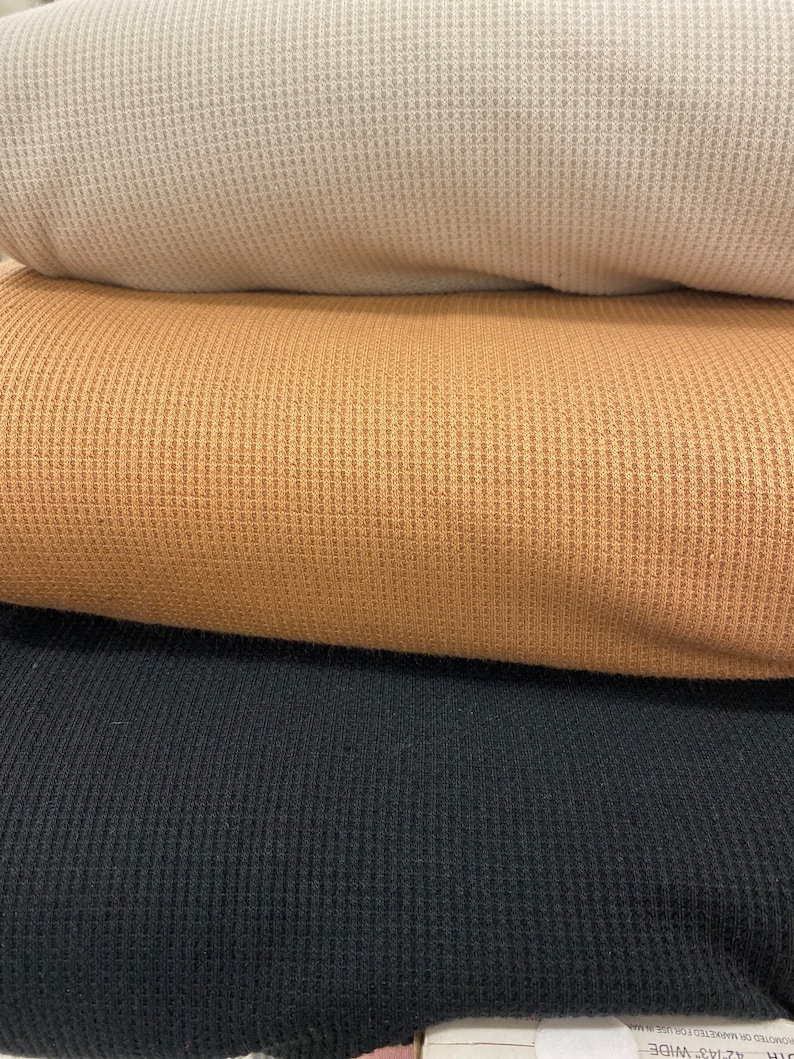 May include: A stack of three folded waffle knit fabrics in neutral colors. The top fabric is a light beige, followed by a tan and then a black fabric. The fabrics have a textured, grid-like pattern.