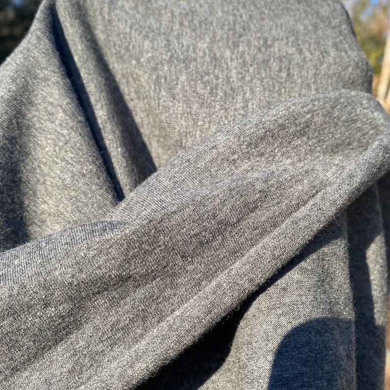 May include: Close-up of a folded section of gray fabric. The material appears to be a soft knit, with visible texture and subtle variations in the gray color. The fabric is draped, creating shadows and highlighting the texture.