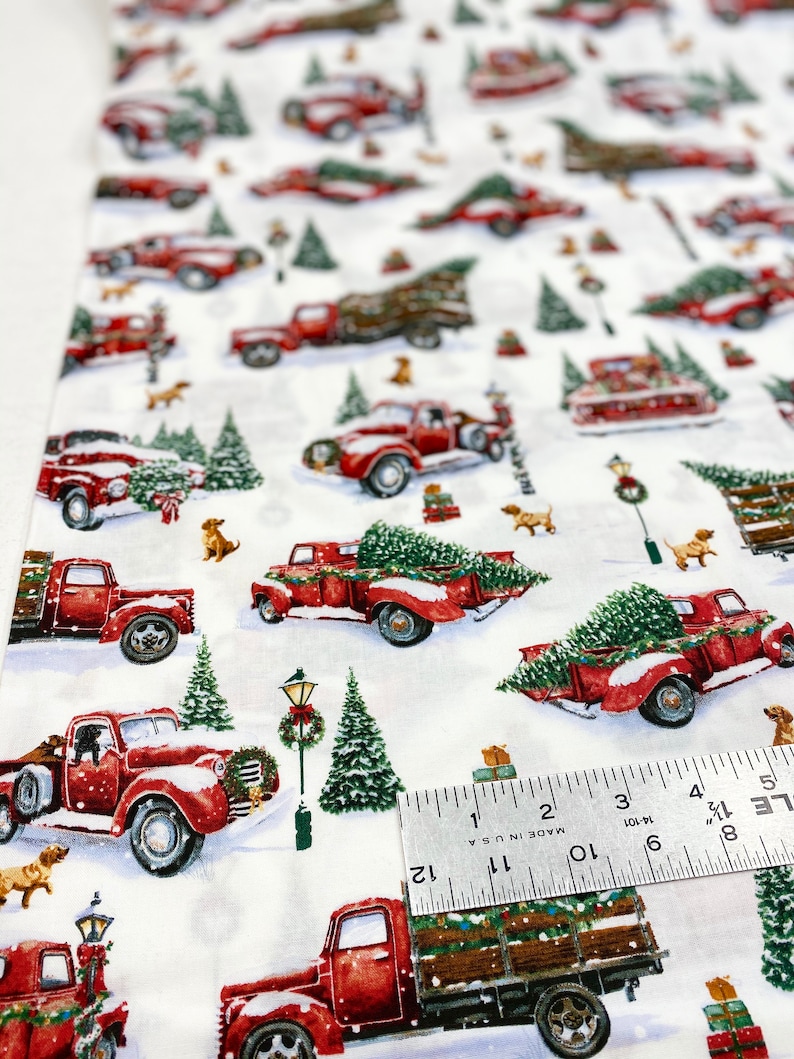K&ouml;nnte beinhalten: Wei&szlig;er Stoff mit einem sich wiederholenden Muster aus roten Oldtimer-Trucks, die Weihnachtsb&auml;ume transportieren, schneebedeckten Tannen und kleinen braunen Hunden. Die Trucks sind mit Schnee bedeckt. Der Stoff wird mit einem Lineal gemessen.
