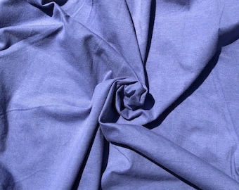 May include: A close-up shot of a piece of fabric in a muted periwinkle color. The fabric is bunched and folded, creating shadows and texture. The material appears soft and flowing, with a slight sheen.