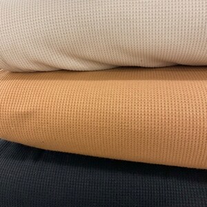 May include: A stack of three waffle-knit fabrics in cream, tan, and black. The close-up shows the textured surface of the material, highlighting the woven pattern. The fabrics are folded, revealing the layers and colors.
