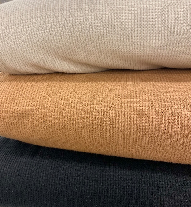 May include: A stack of three waffle-knit fabrics in cream, tan, and black. The close-up shows the textured surface of the material, highlighting the woven pattern. The fabrics are folded, revealing the layers and colors.