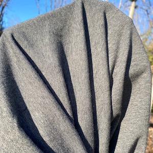 May include: Close-up of a gray fabric with visible texture and folds. The fabric is a solid, medium gray color, with shadows creating vertical lines. The material appears to be soft and possibly a knit or woven textile.