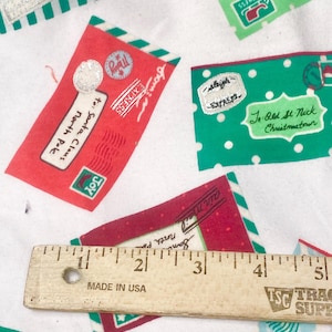 May include: White fabric with a repeating pattern of Christmas-themed envelopes in red, green, and blue. The envelopes have text such as "To: Santa Claus, North Pole" and "Express". A wooden ruler with "Made in USA" is also visible.